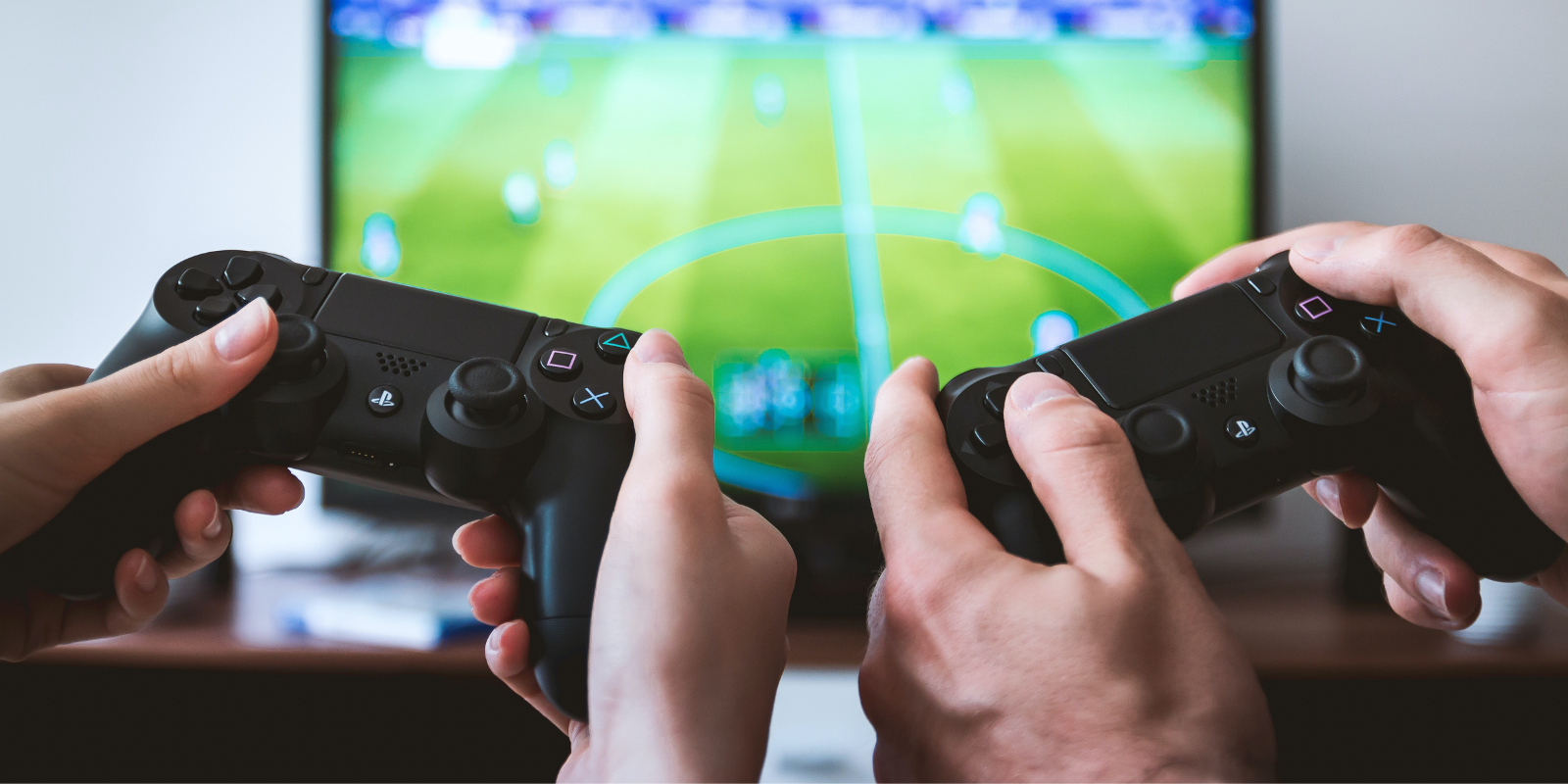 Two individuals holding video game controllers, the colour of the controllers are black, they are engrossed in a football game, in front of a large plasma television screen. You can't see their faces, only their hands with the controllers. 