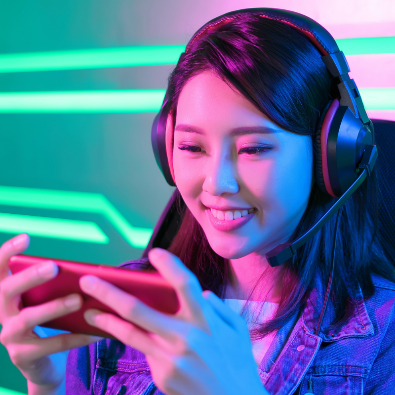 A smiling woman with black hair wears pink headphones, looking at her phone while playing a game. Neon lights illuminate the background.