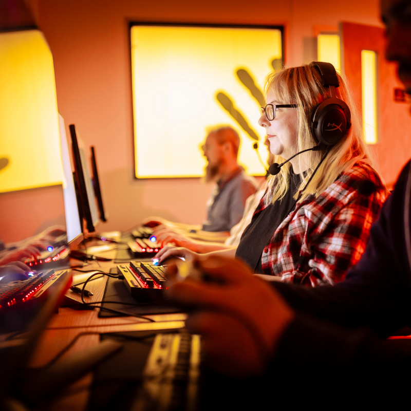 Jen, Will, and Mattie, colleagues at Kwalee, engage in gaming on PCs within a warmly illuminated red room. The atmosphere suggests camaraderie and enjoyment as they immerse themselves in gameplay, fostering team bonding and relaxation.