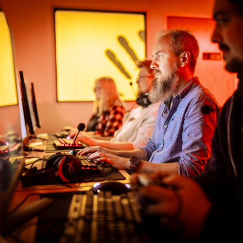 A cohesive group of people diligently at work on their computers, sitting together at a table. A united and focused team. They are all sat playing video games. There is a yellow picture in the background of the Kwalee logo, which is a handprint. A productive scene.  