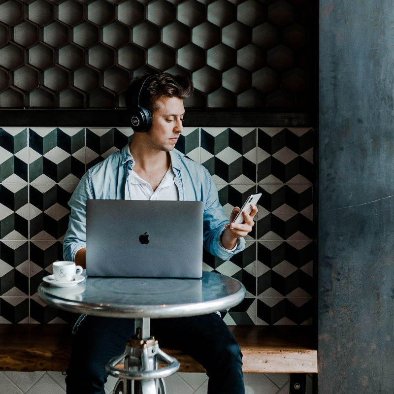 The photo shows a man wearing wireless black headphones, staring down at his white iphone. In front of him on a table is a silver mac with the apple logo showing, and a coffee cup. The scenery is that he could be in a cafe doing some work, researching, or content creating, 