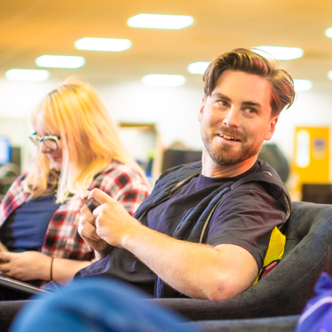 An individual, Head of Development Tom Bromwich, is seated comfortably on a couch, in his hands are his phone which he is playing a mobile game, his face is facing the camera, possibly engaged in leisure or contemplation